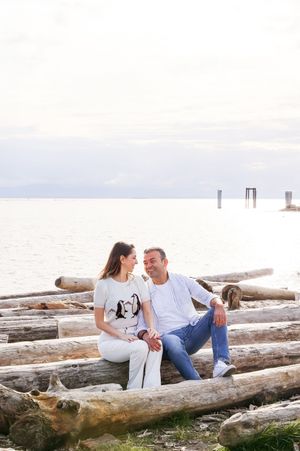 A couple sits together on driftwood logs by the water during a romantic sunset photoshoot at the beach.