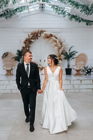 Couple walks hand in hand in formal wedding attire under a decorated white ceiling.
