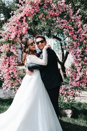 A couple in wedding attire embrace under a stunning arch of pink cherry blossoms in a garden setting.