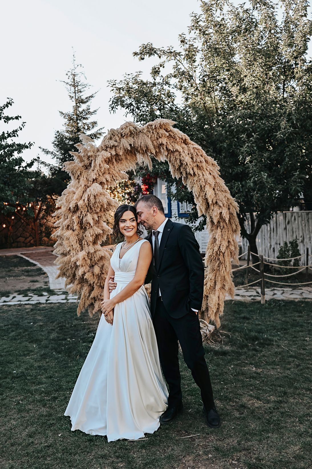 Wedding couple poses under a rustic brown arch with white dress and black suit.