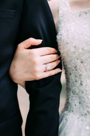 Close-up of a hand wearing an engagement ring against a black suit and white beaded dress.