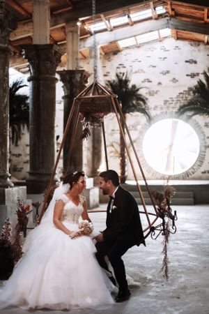 Romantic wedding moment captured under an ornate hanging swing in a columned venue.