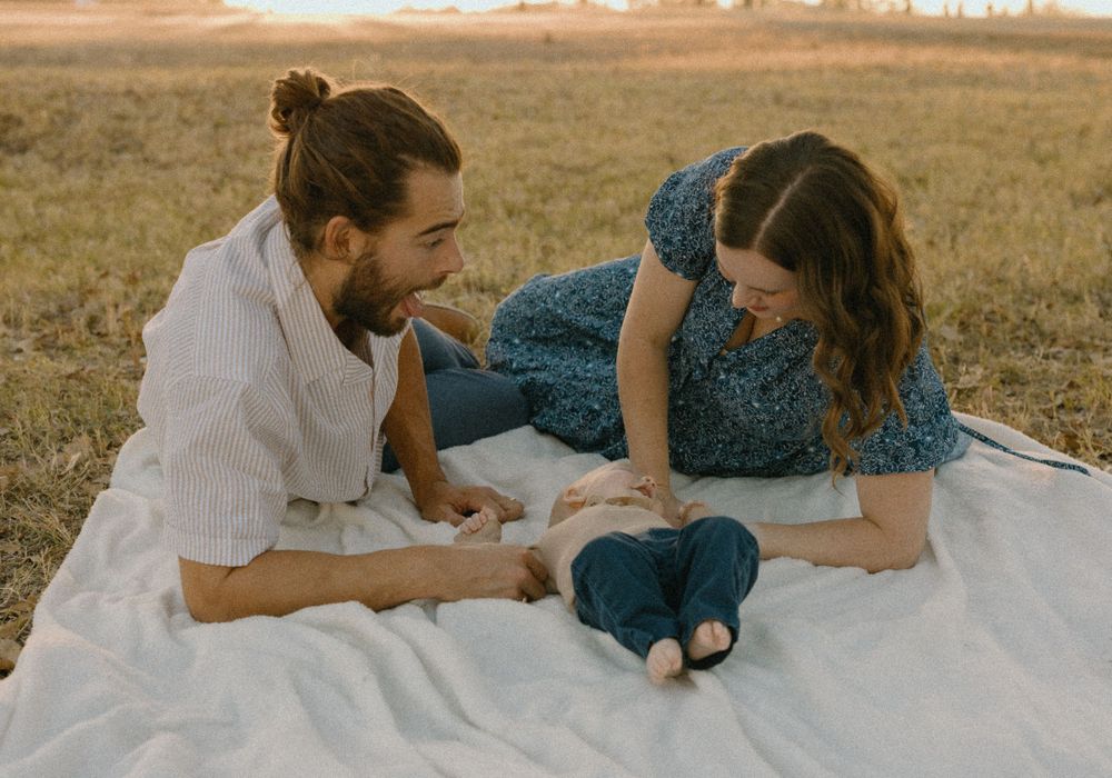 a couple laying on a white blanket in a field looking at their baby joyfully