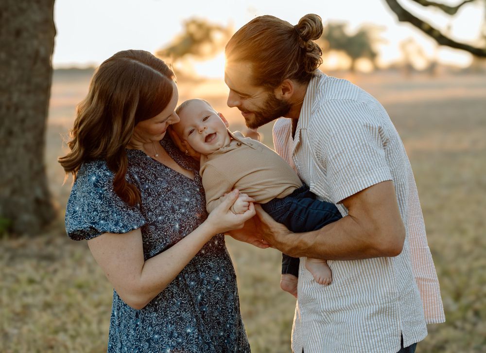 A couple embraces their laughing baby during a sunset photo session in a field.