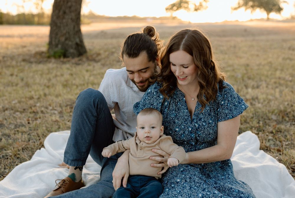 A couple sits on a white blanket with their baby during a golden hour photoshoot in a field.