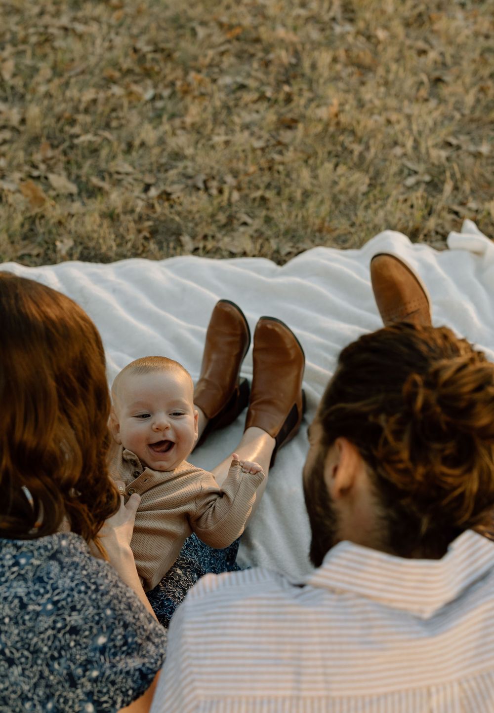family photo of a mom, dad, and baby on a white blanket in a field