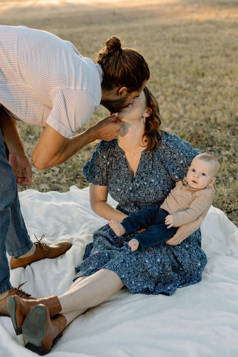 family photo of a mom, dad, and baby on a white blanket in a field