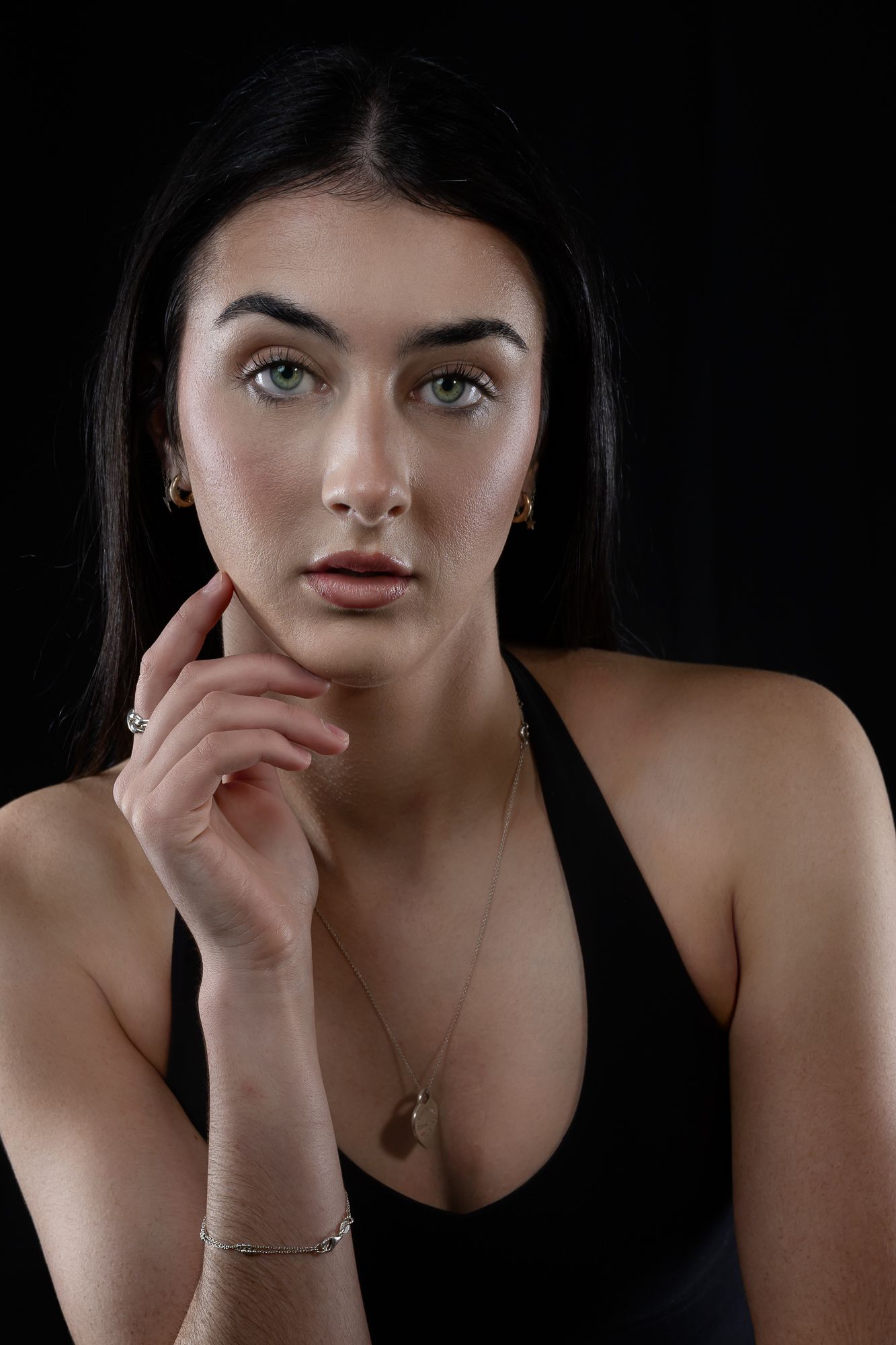 Dramatic portrait in black top with delicate jewelry and thoughtful pose against dark backdrop.