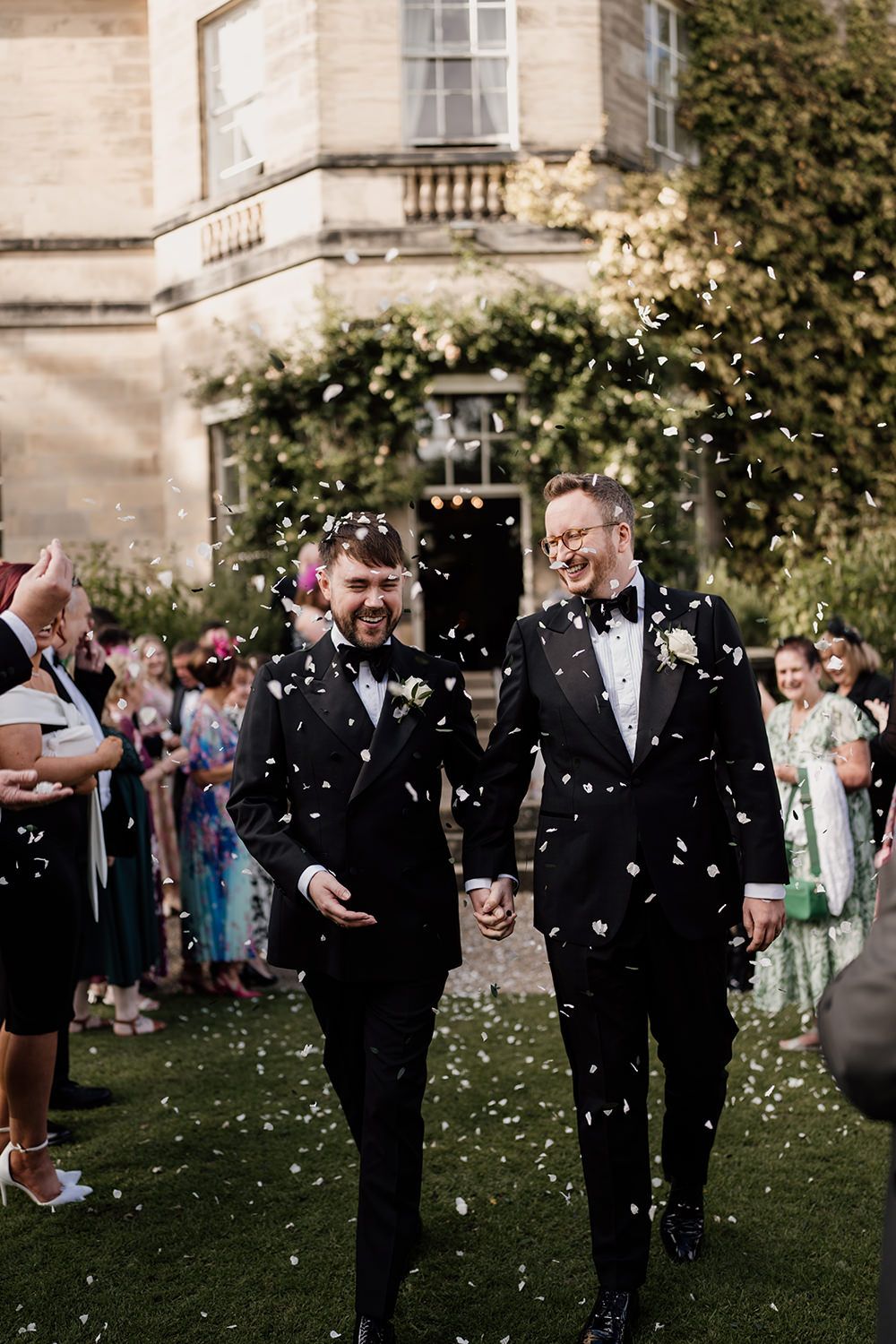 Two grooms walking holding hands while guests throw confetti at Middleton Lodge Estate Main House.