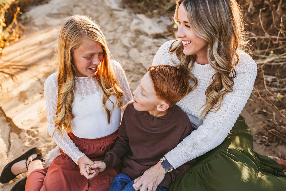 A mother smiles warmly with her two children, a girl and boy, sitting together outdoors in golden light.