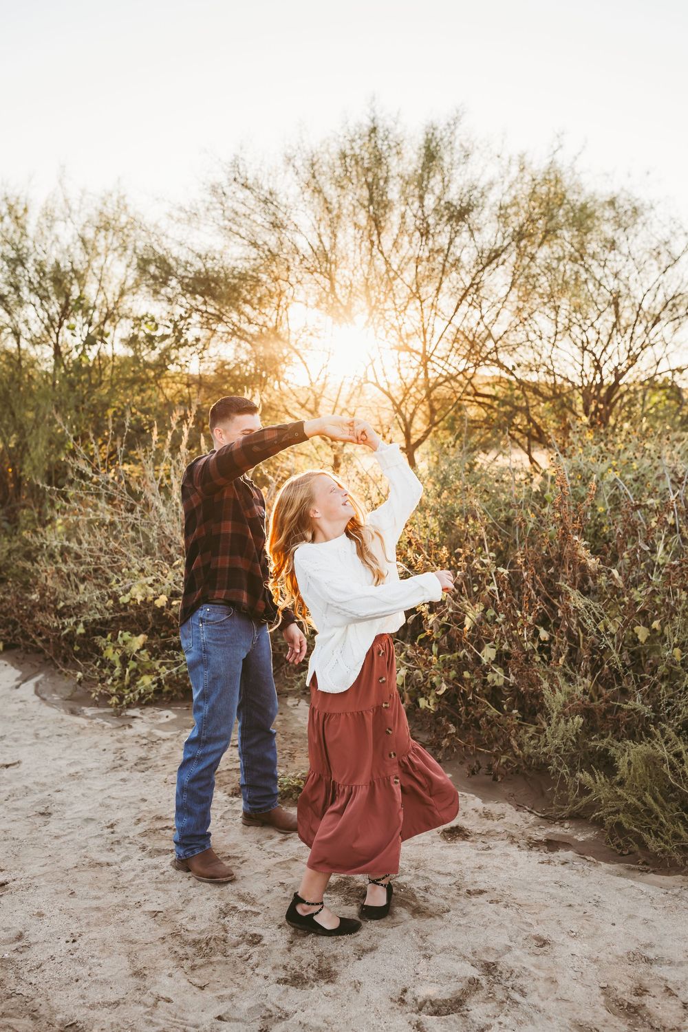 A couple dances outdoors at golden hour, woman in red skirt twirling, surrounded by desert shrubs in warm sunset light.