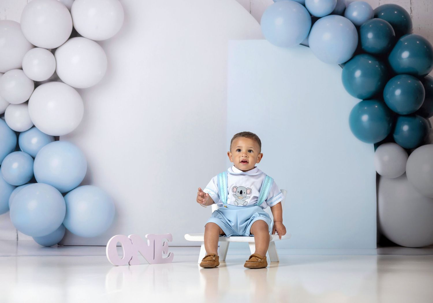 Baby boy in blue romper sits before a balloon arch display with ONE sign for a first birthday photo shoot.