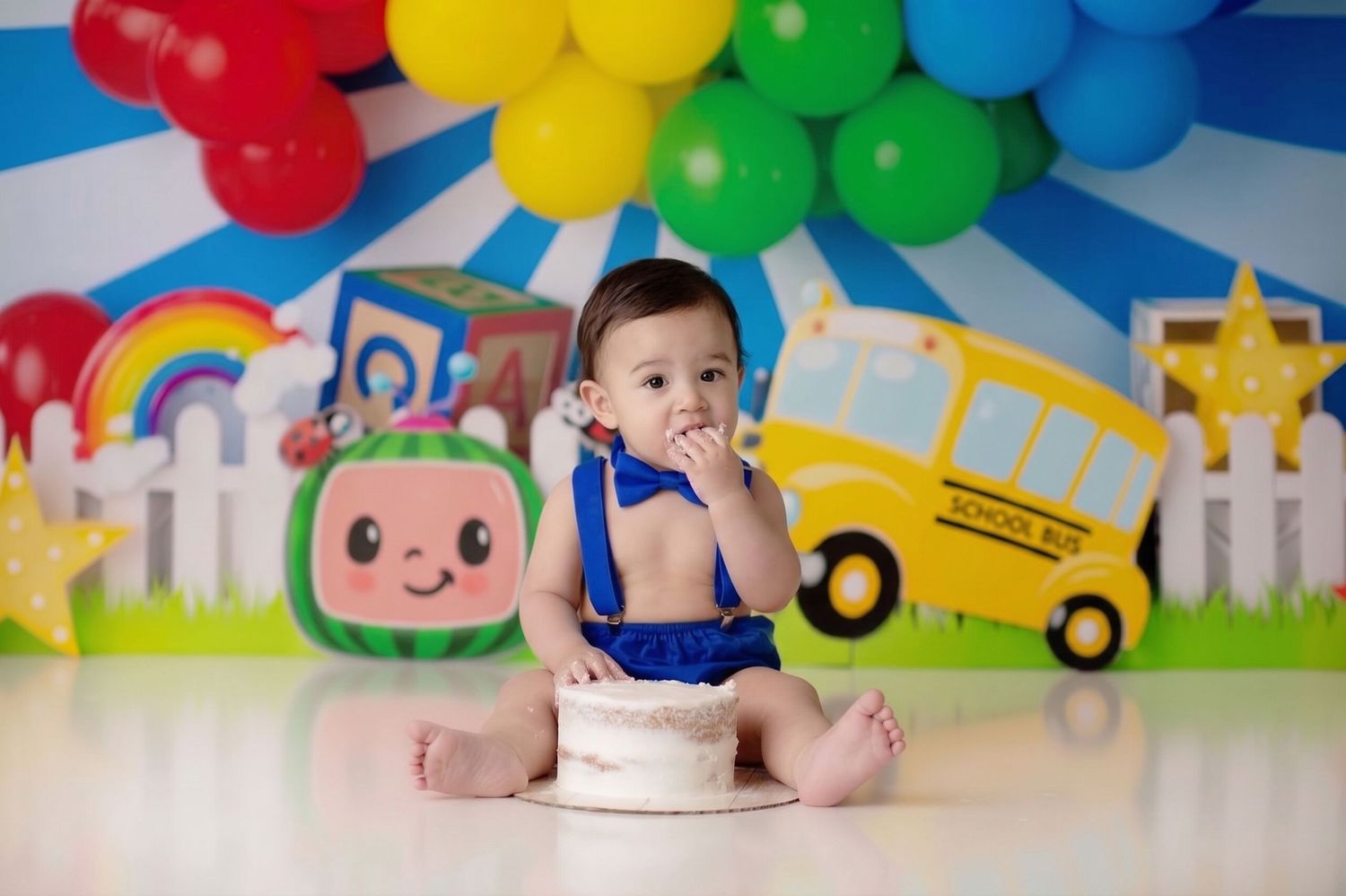 Baby boy in blue romper smashes first birthday cake at Cocomelon themed photo shoot with colorful balloons.