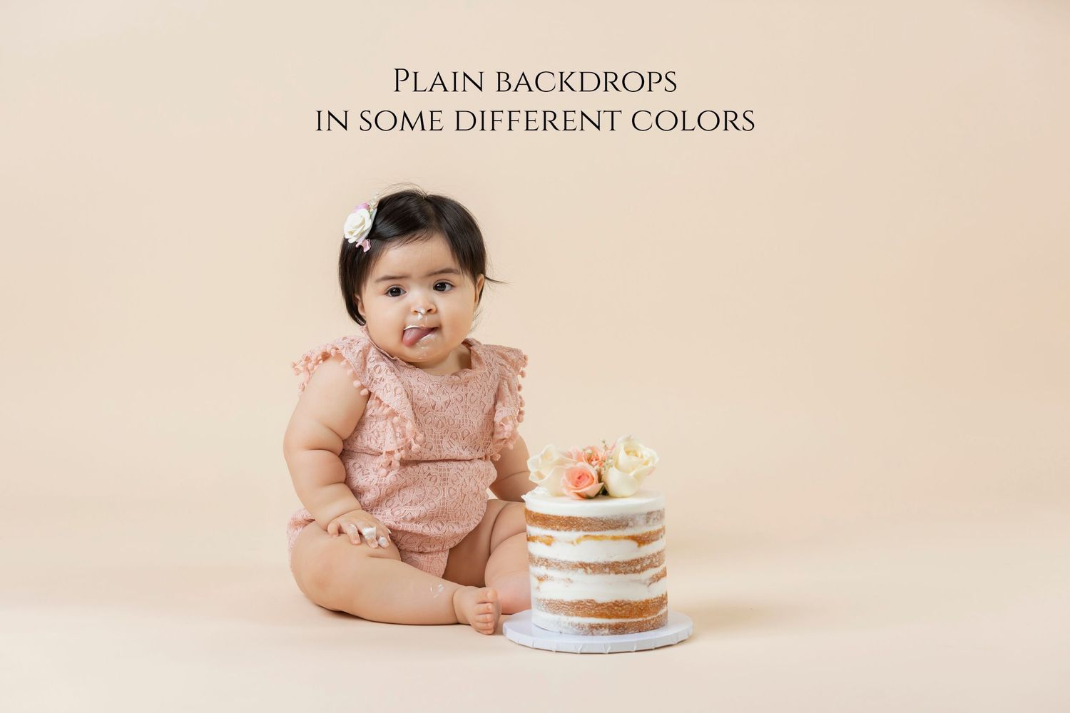 Baby girl in pink romper sits beside a small naked cake on a plain beige backdrop for a cake smash photo.