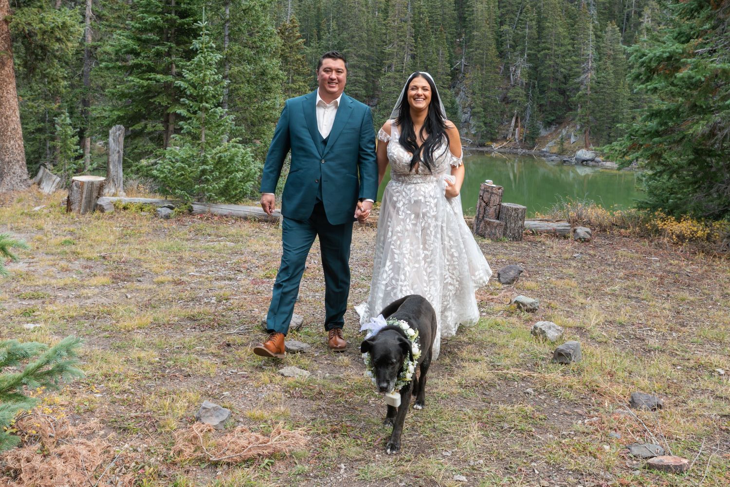 An eloping couple and their dog walk back down the aisle after saying vows with an alpine lake and pine forest behind them. Both are smiling