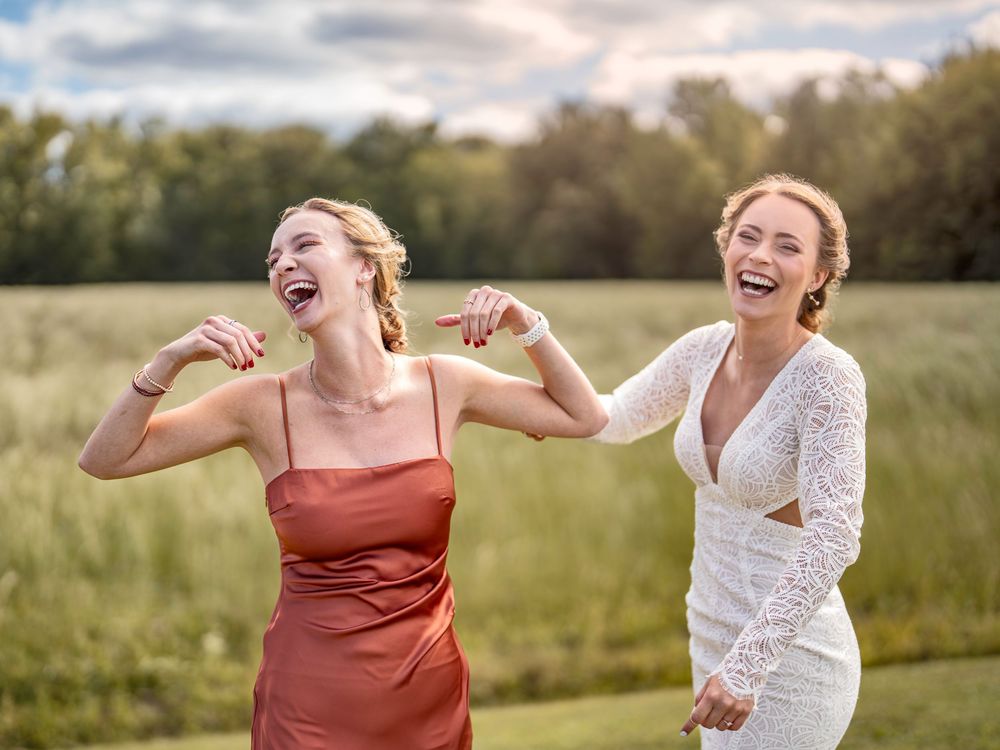 Bridal party members in white dresses and rust colored gowns laugh joyfully together in a golden sunlit field.