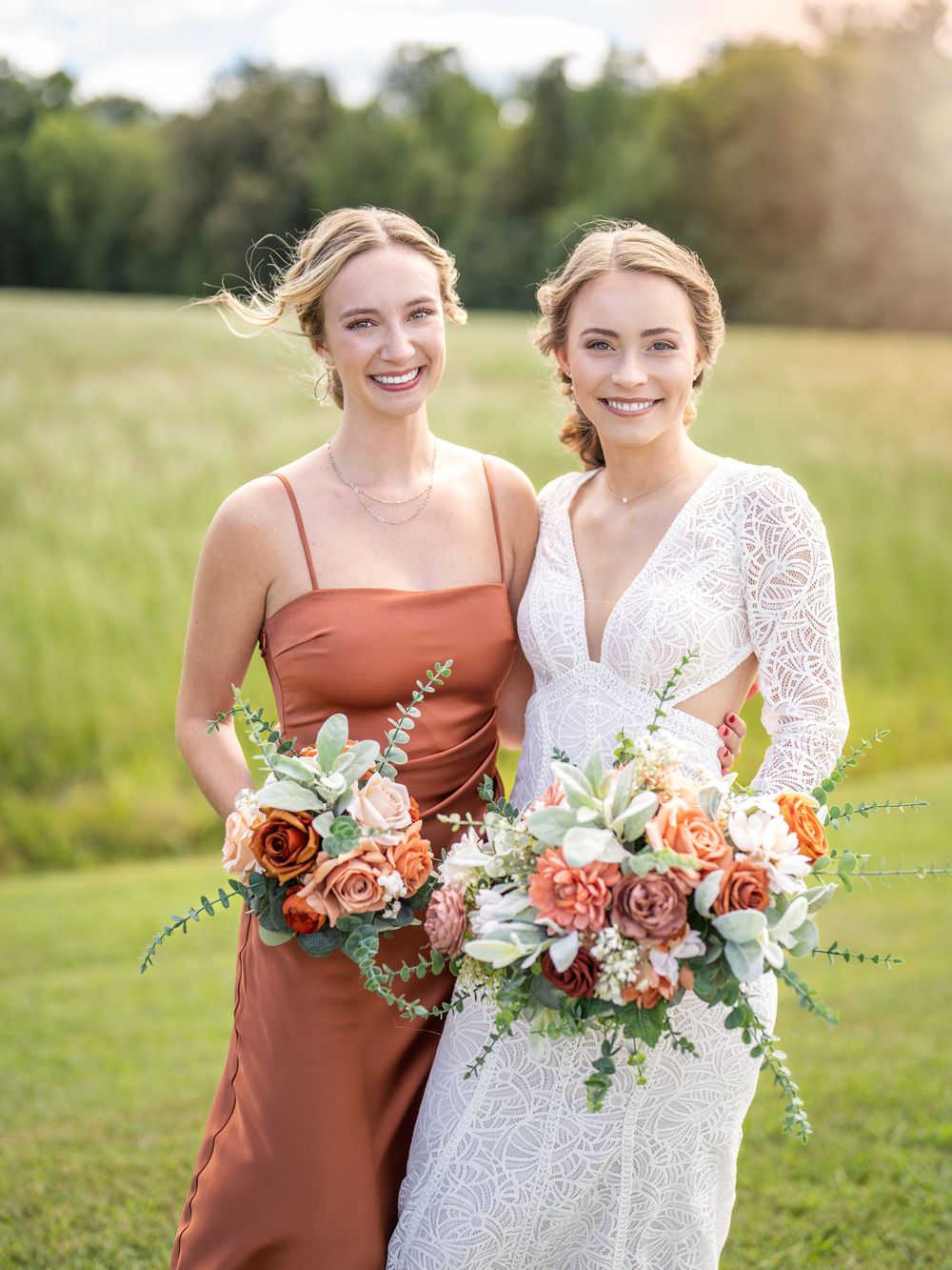 Two people wearing rust-colored and white dresses embrace while holding coral and peach floral bouquets in a sunlit field.