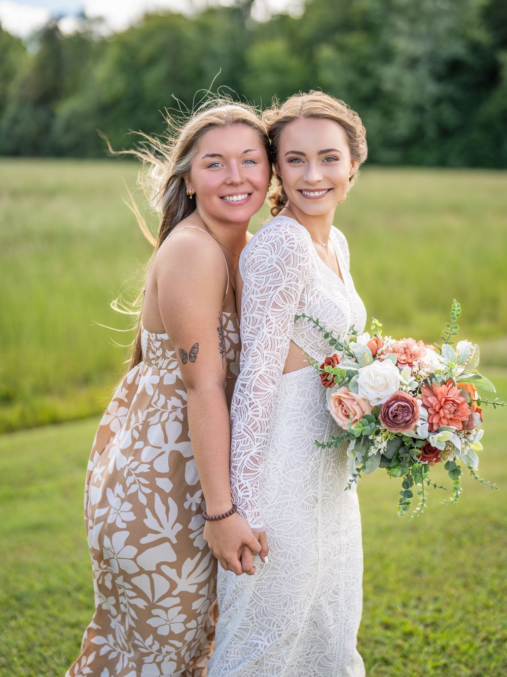 Two people in dresses stand together on grassy field holding colorful floral bouquets.