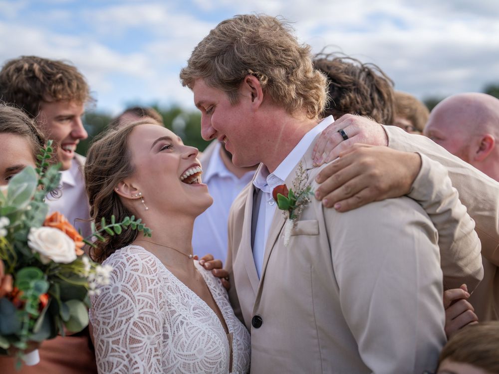 A joyful wedding moment captured outdoors as newlyweds share a laugh surrounded by their wedding party under cloudy skies.