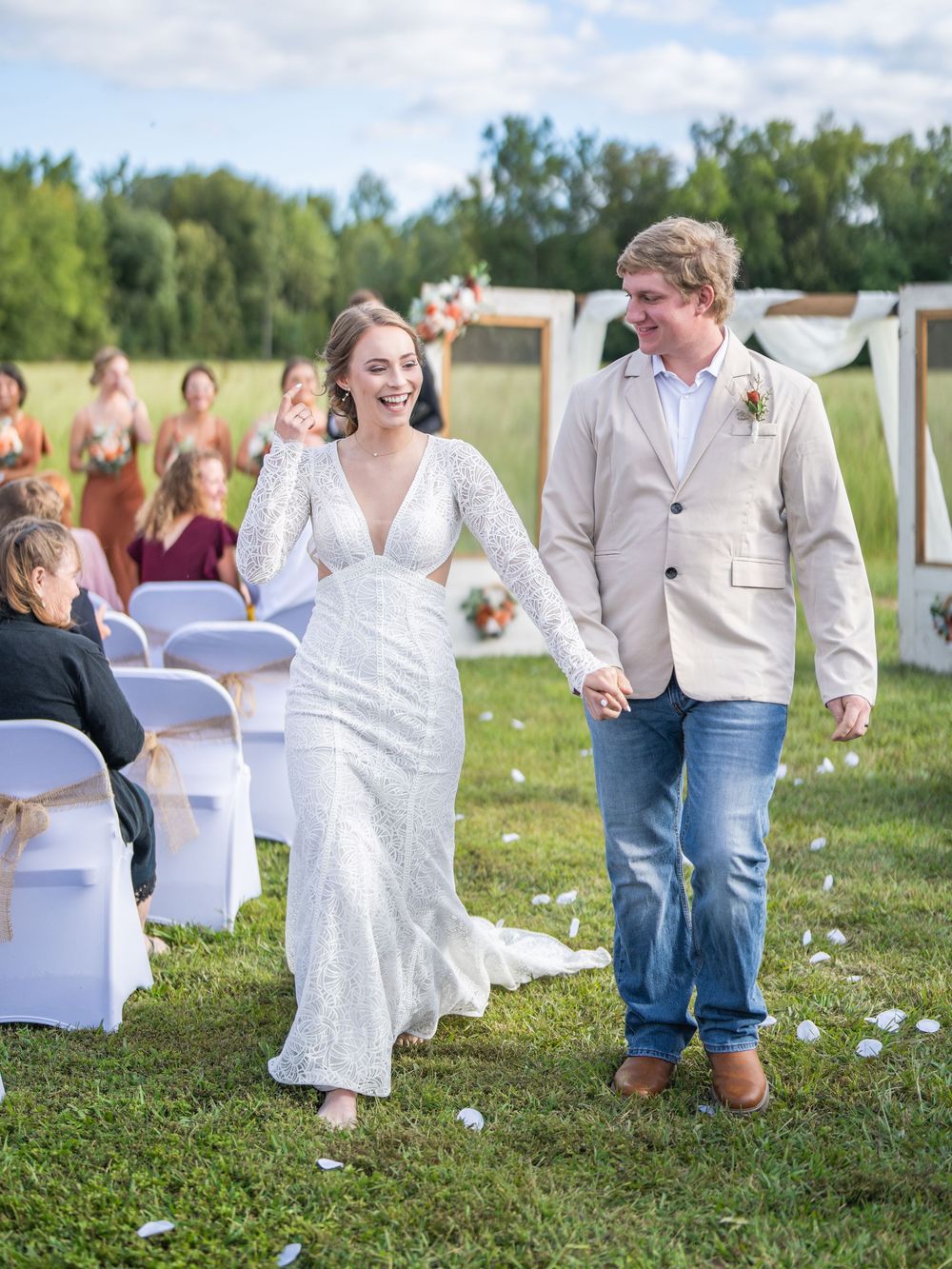A joyful couple walks down the aisle at an outdoor wedding ceremony with white chairs and greenery in the background.