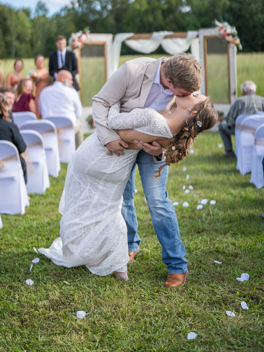 A couple in wedding attire shares a fun dip pose at their outdoor ceremony with white chairs and trees in the background.