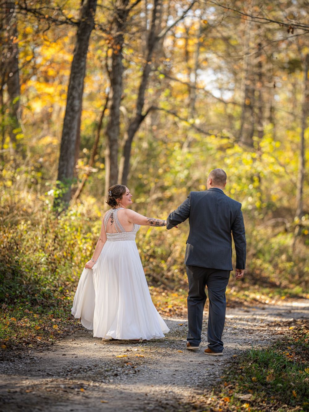 A couple walks together on a woodland path surrounded by golden fall foliage and warm autumn sunlight.