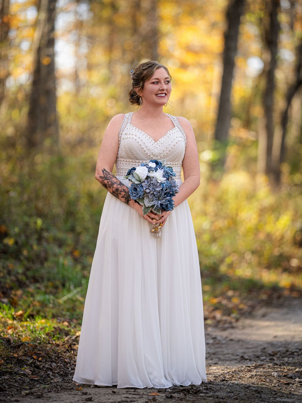 A person in a white flowing wedding dress poses on a forest path with golden autumn foliage in the background.