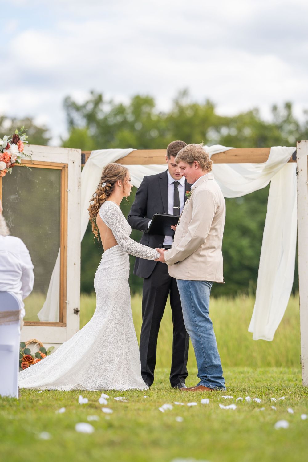 A rustic outdoor wedding ceremony takes place in a grassy field with a white fabric draped arch and vintage mirror decor.