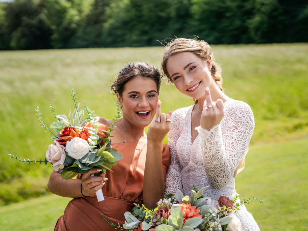 Two figures in wedding attire laugh joyfully while holding vibrant floral bouquets in a sunny meadow.