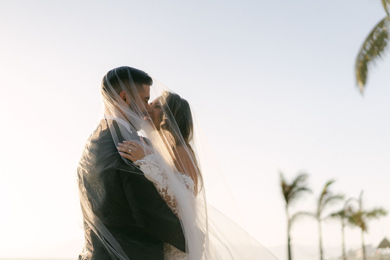 Newlyweds share a romantic kiss under a flowing veil at sunset.