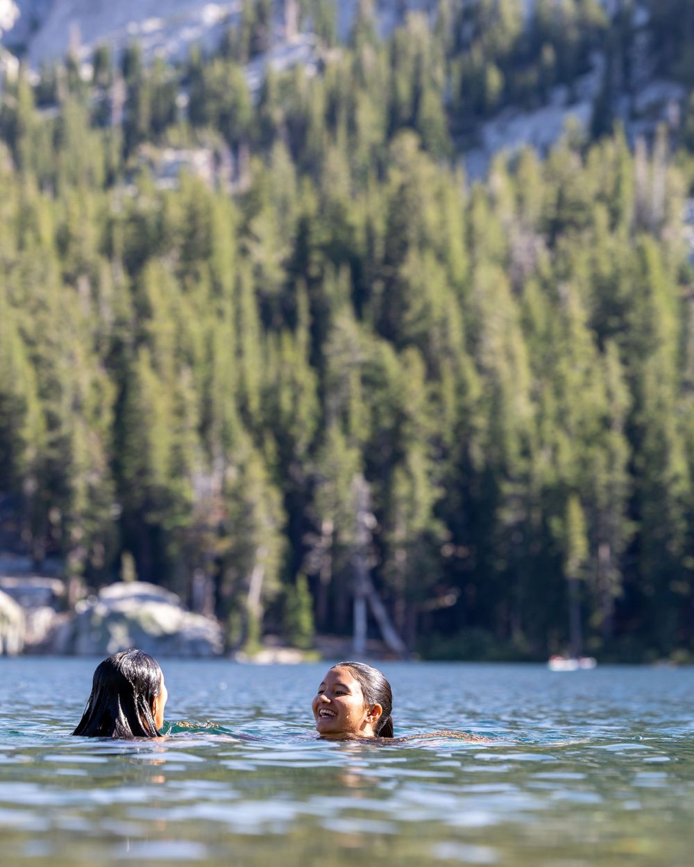 Two people swimming in a serene mountain lake surrounded by tall pine trees and rocky cliffs.