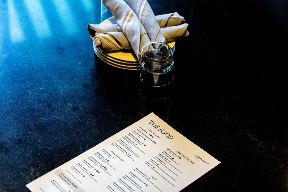A restaurant menu titled The Food lies on a dark table beside a folded napkin on a plate and a glass.