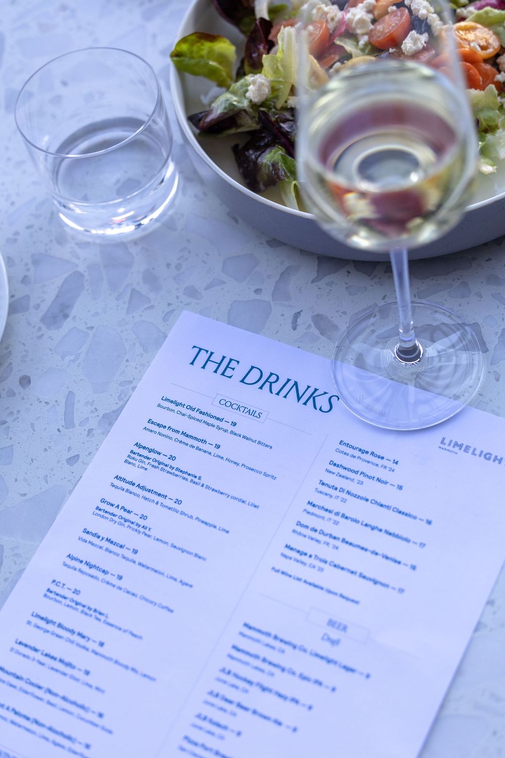 A drinks menu beside a wine glass and fresh salad at Limelight restaurant on a marble table.