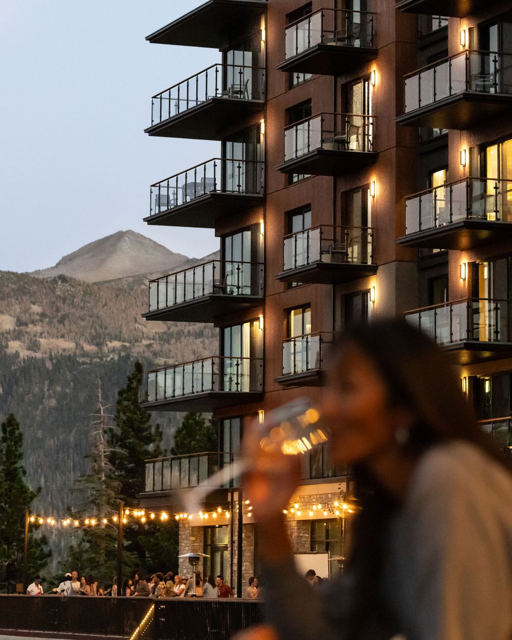 A woman holds a wine glass on a mountain resort terrace at dusk, with a lit high-rise hotel and peaks in the background.