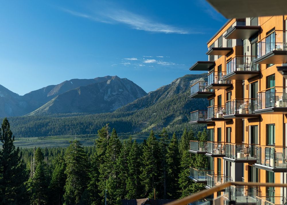 Modern hotel balconies overlooking lush pine forest and dramatic mountain peaks under a clear blue sky.