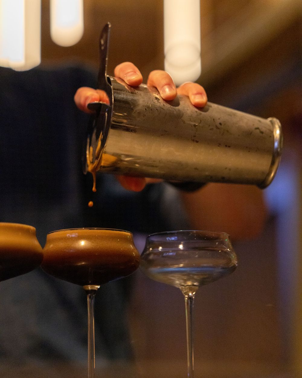 A bartender pours a dark cocktail from a shaker into elegant coupe glasses at a bar.