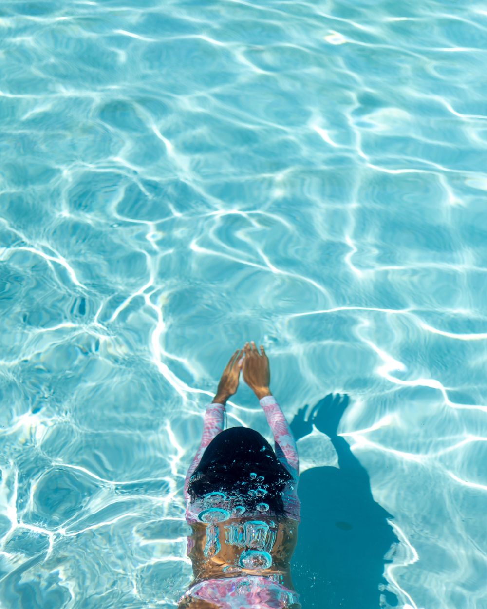 Aerial view of child in pink swimsuit diving into crystal clear turquoise swimming pool.