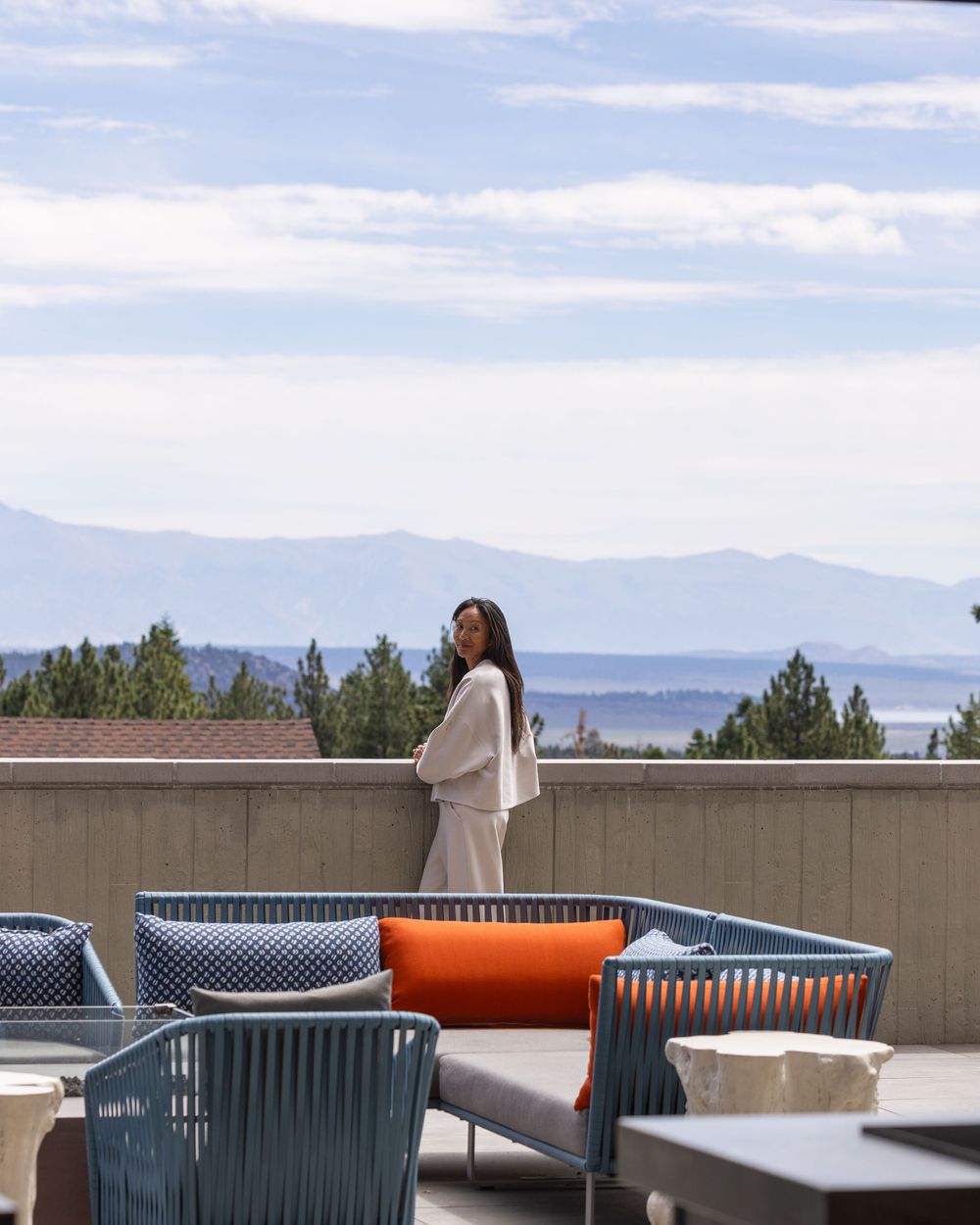 Woman in beige outfit stands on hotel terrace with mountain and lake views under a cloudy sky.