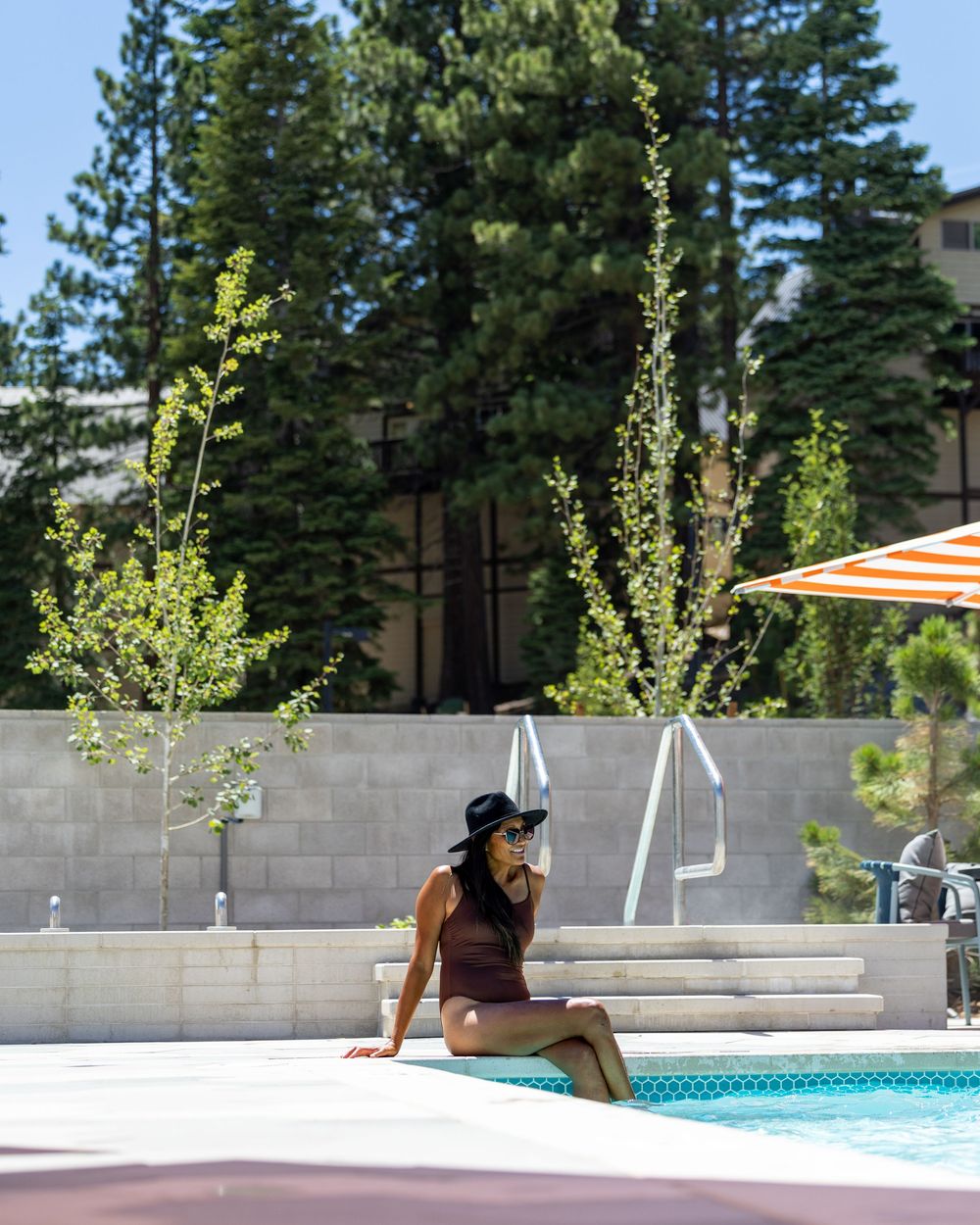 Woman in burgundy swimsuit and black hat relaxing by a sunny outdoor pool surrounded by tall pine trees.