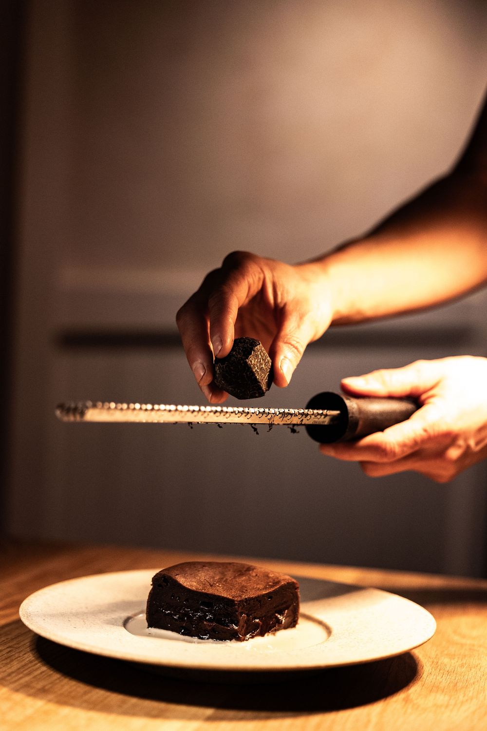 Chef grating dark chocolate over a rich chocolate brownie on a white plate in a moody restaurant setting.