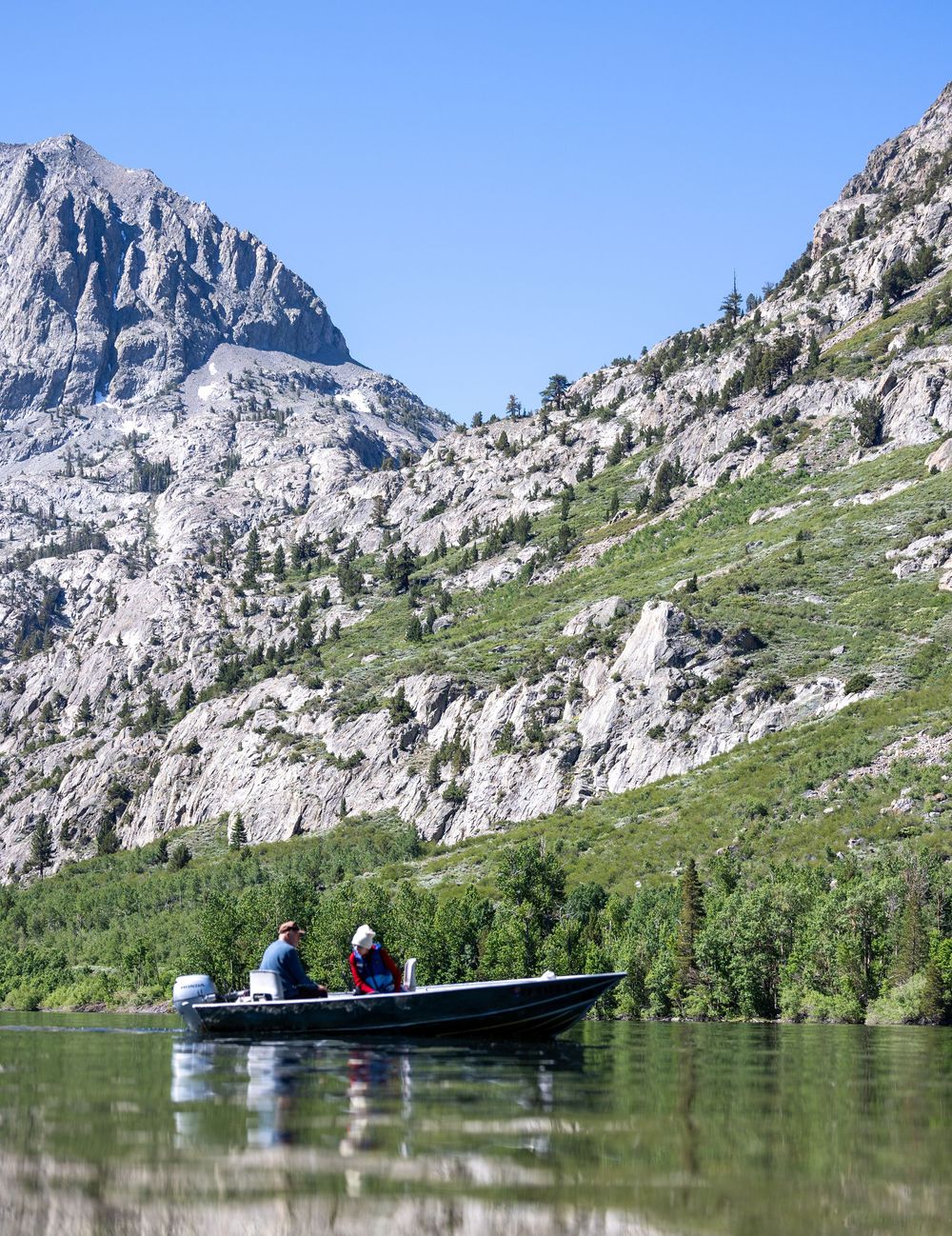 Two anglers fishing from a motorboat on a serene alpine lake surrounded by rocky mountains and pine trees.