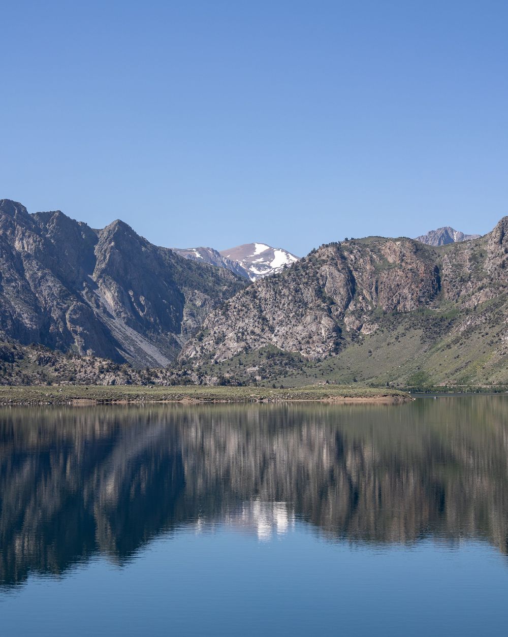 Snow-capped mountain peaks reflect perfectly in a calm alpine lake surrounded by rugged terrain under clear blue skies.