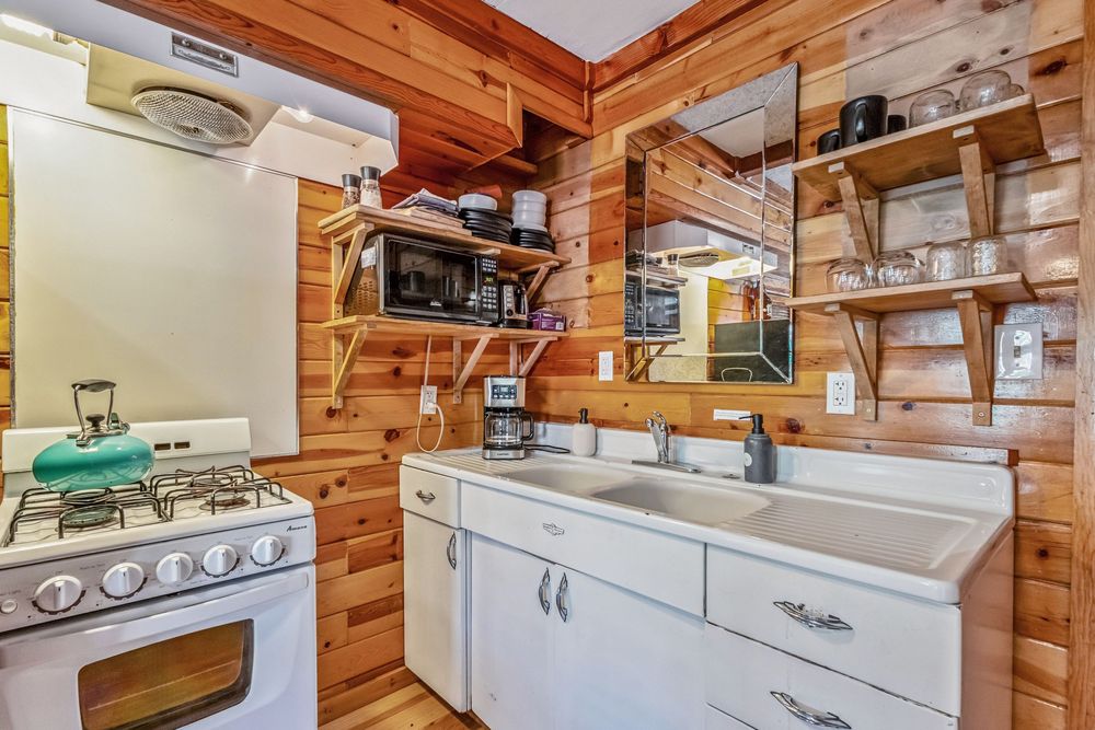 Rustic wood-paneled cabin kitchen with white gas stove, double sink vanity, and open shelving with small appliances.