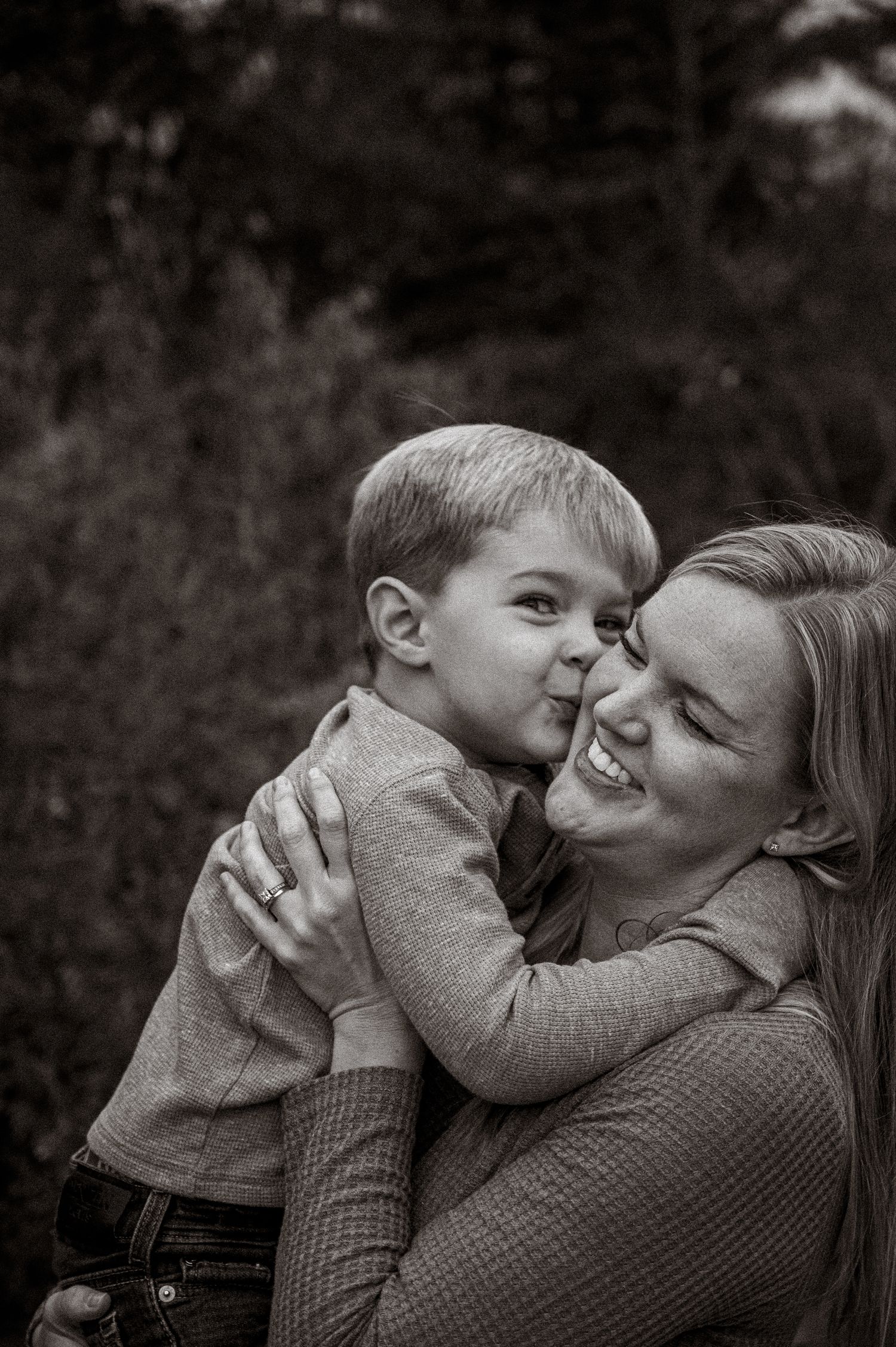 Black and white photo of a young boy giving his smiling mother a kiss on the cheek outdoors.