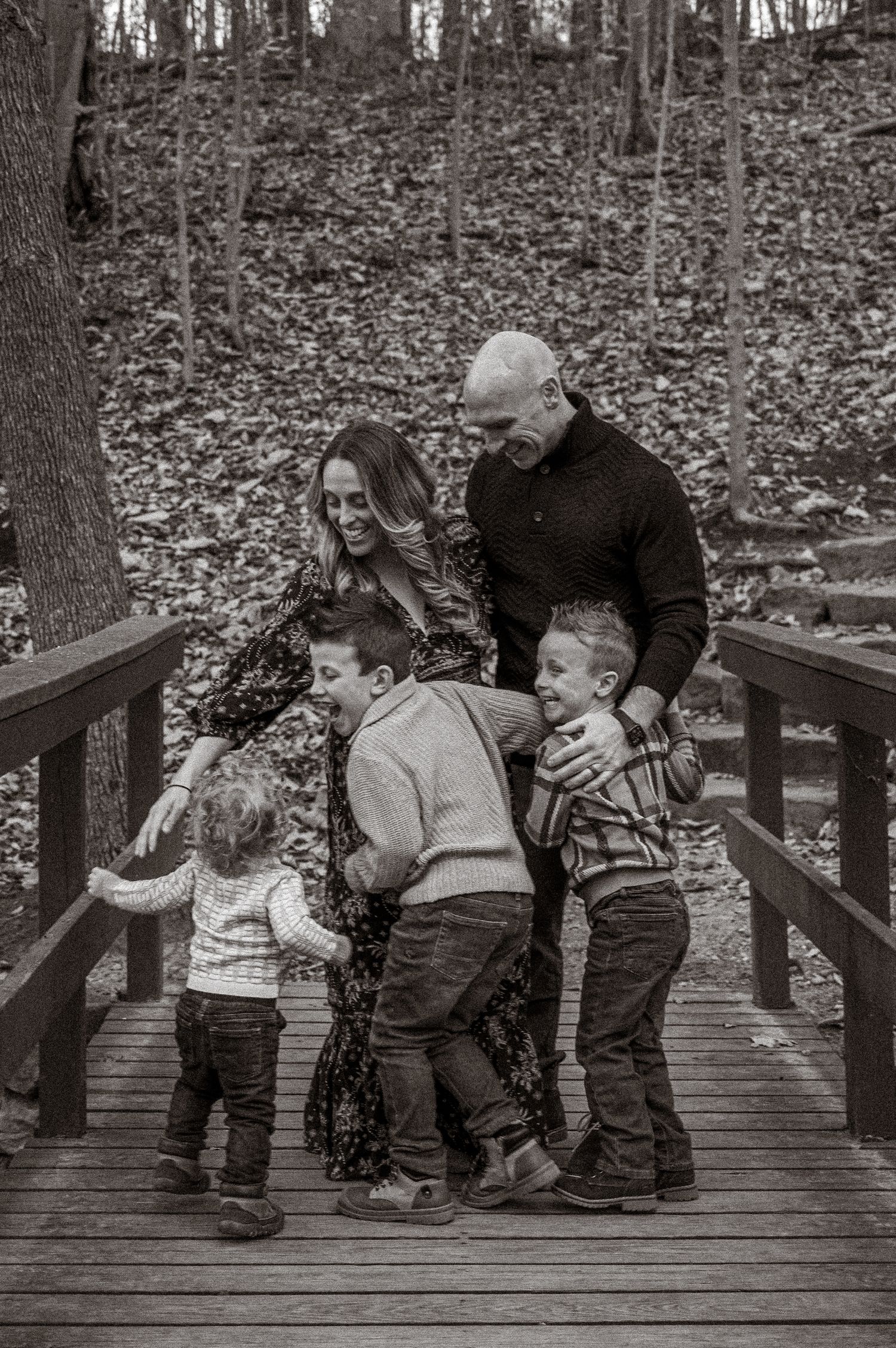 Black and white family portrait of parents and three children laughing on a wooden bridge.