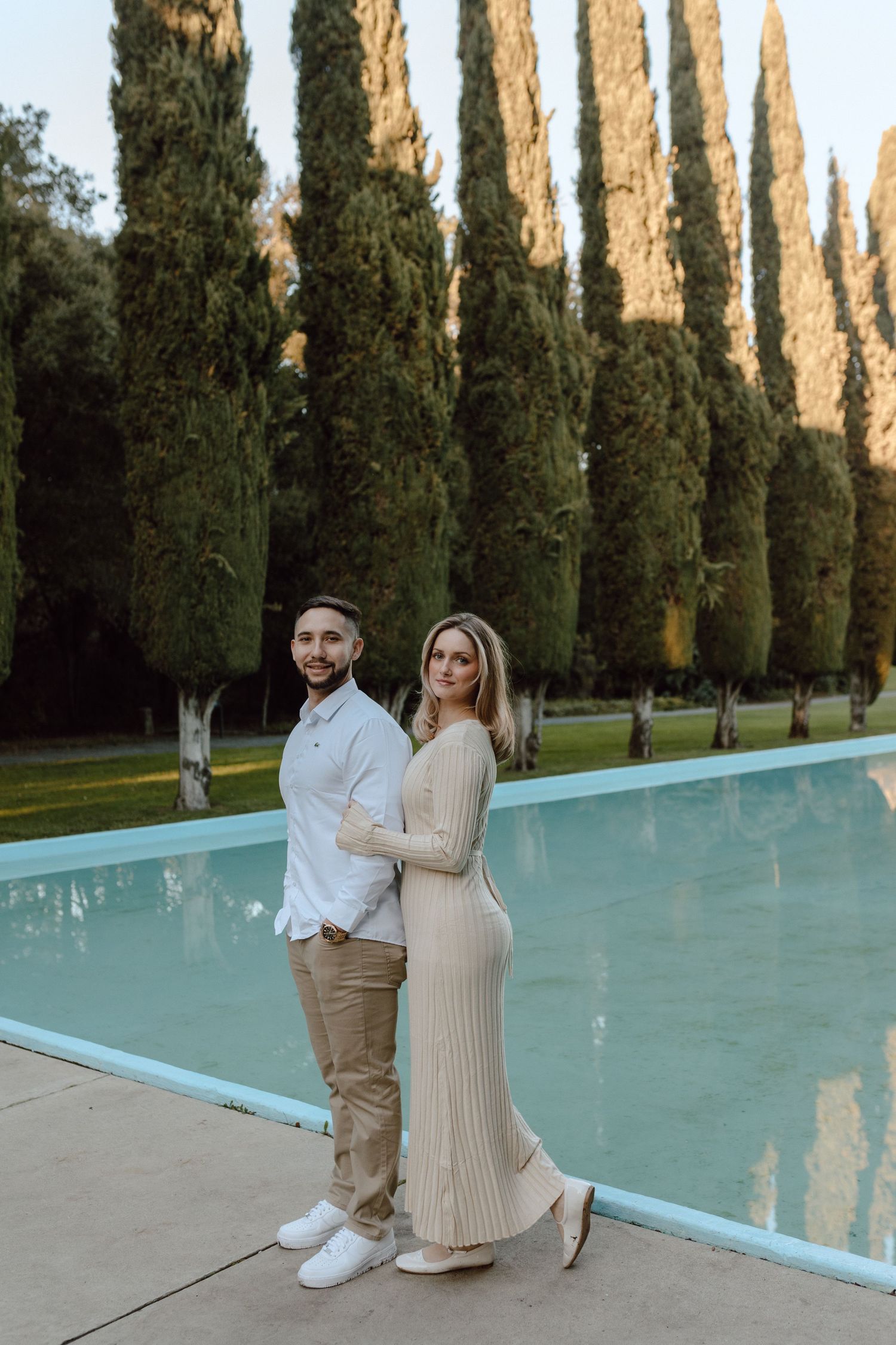 Couple posing by swimming pool with row of tall cypress trees in background.