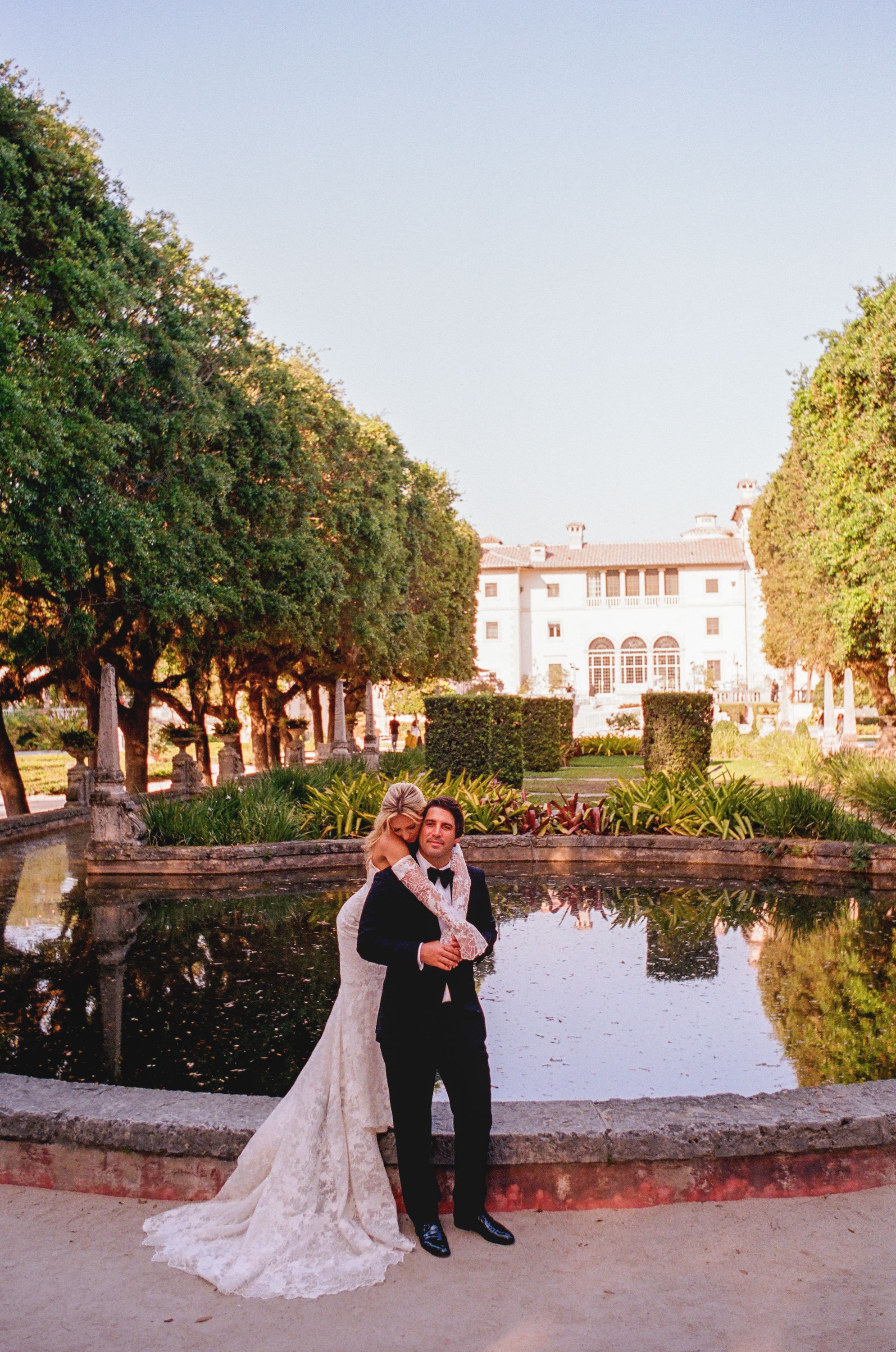 Romantic wedding couple standing by reflective pond at sunset in a lush garden setting with classic architecture backdrop.