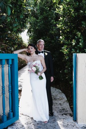 Bride in white gown holding pink bouquet stands with groom in black tuxedo by blue gate surrounded by lush greenery.