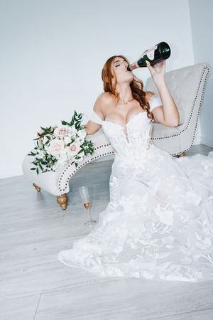 Bride in white lace gown drinking champagne from bottle, seated by floral bouquet on elegant sofa.