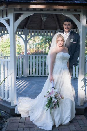 Bride in white gown holding bouquet stands with groom in gray suit under a white gazebo outdoors.